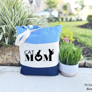 Puede incluir: Una bolsa de tela azul, blanca y azul marino con el texto "CAT MOM" y dos siluetas de gatos negros. La bolsa tiene dos asas blancas.