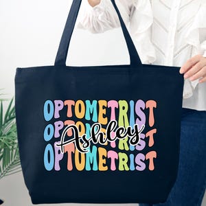 May include: A navy blue tote bag with the word "OPTOMETRIST" in colorful, bubble letters. The name "Ashley" is written in cursive script in the center of the bag. The bag has black handles.