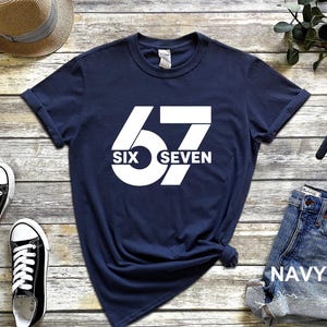 May include: Navy blue t-shirt with a large white graphic of the number "67" and the words "SIX" and "SEVEN" in a bold, sans-serif font. The shirt is styled with rolled-up sleeves.