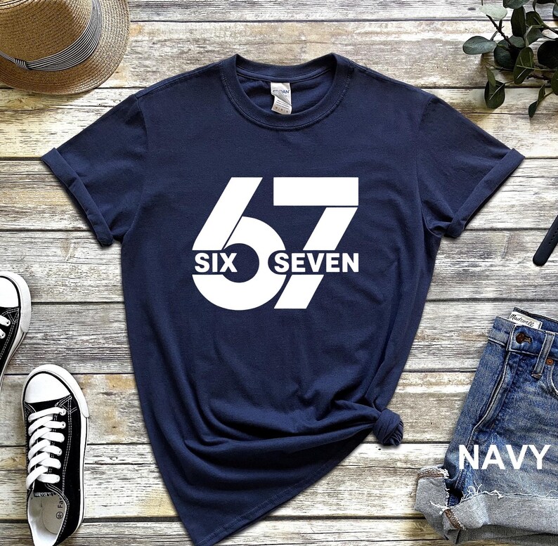 May include: Navy blue t-shirt with a large white graphic of the number "67" and the words "SIX" and "SEVEN" in a bold, sans-serif font. The shirt is styled with rolled-up sleeves.