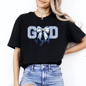 K&ouml;nnte beinhalten: Schwarzes T-Shirt mit einem blauen und wei&szlig;en Blumenmuster, auf dem "GOD is good" steht, mit einer blauen und wei&szlig;en Schleife in der Mitte des Wortes "GOD".