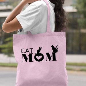 Puede incluir: Una bolsa de tela de color rosa claro con asas negras y el texto "CAT MOM" con dos siluetas de gatos negros.