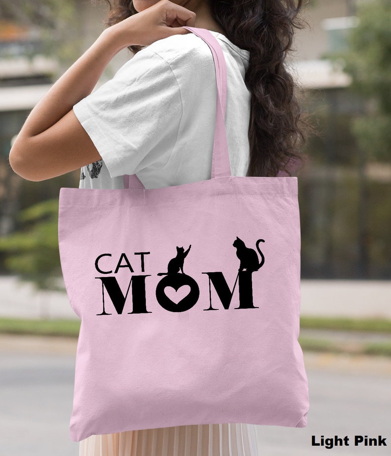 Puede incluir: Una bolsa de tela de color rosa claro con asas negras y el texto "CAT MOM" con dos siluetas de gatos negros.