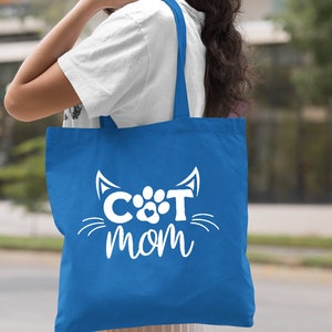 Pode incluir: Uma bolsa tote azul royal com texto branco que diz "CAT MOM" e um rosto de gato branco com uma pegada no meio.