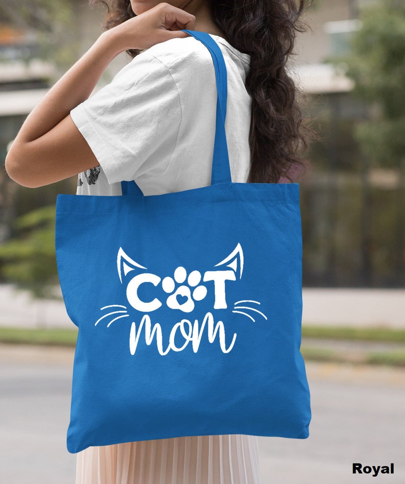 Pode incluir: Uma bolsa tote azul royal com texto branco que diz "CAT MOM" e um rosto de gato branco com uma pegada no meio.