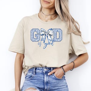 K&ouml;nnte beinhalten: Ein beiges T-Shirt mit einem blauen und wei&szlig;en Blumendesign, das "GOD is good" buchstabiert, mit einer blauen Schleife in der Mitte des Wortes "GOD".