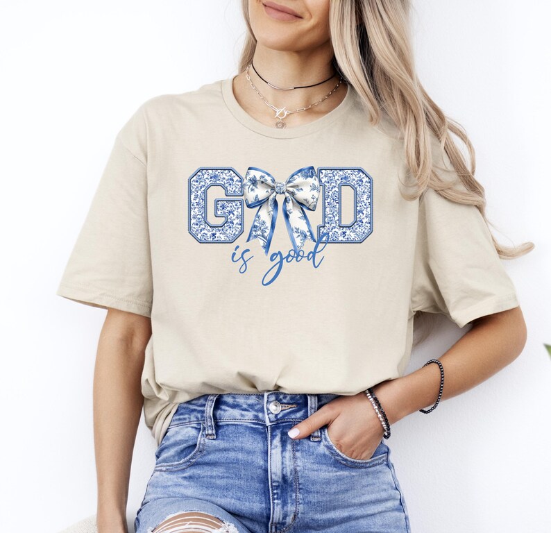 K&ouml;nnte beinhalten: Ein beiges T-Shirt mit einem blauen und wei&szlig;en Blumendesign, das "GOD is good" buchstabiert, mit einer blauen Schleife in der Mitte des Wortes "GOD".