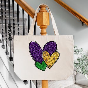Puede incluir: Una bolsa de lona blanca con tres corazones de purpurina. Los corazones son morados, dorados y verdes. La bolsa tiene un asa larga.