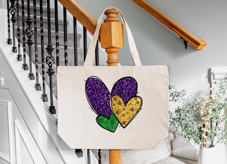 Puede incluir: Una bolsa de lona blanca con tres corazones de purpurina. Los corazones son morados, dorados y verdes. La bolsa tiene un asa larga.