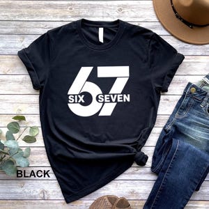 May include: Black t-shirt with a large white graphic of the number 67. The word "SIX" is centered below the 6 and the word "SEVEN" is centered below the 7. The word "BLACK" is printed at the bottom left of the shirt.
