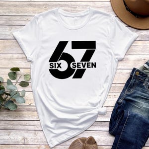 May include: White t-shirt with a black graphic design. The design features the number "67" with the words "SIX" and "SEVEN" incorporated into the design. The shirt is made of a soft material.