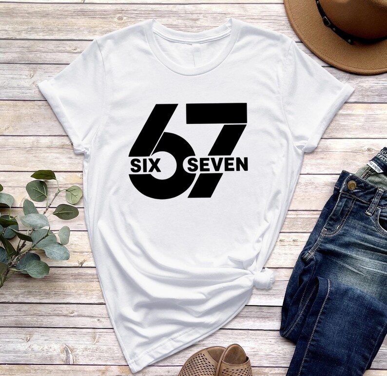 May include: White t-shirt with a black graphic design. The design features the number "67" with the words "SIX" and "SEVEN" incorporated into the design. The shirt is made of a soft material.