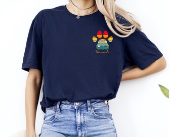 May include: Navy blue t-shirt featuring a paw print design with a stethoscope and the name "Sarah" in a retro style. The paw print is in shades of orange, yellow, and green. The shirt is a casual style.