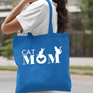 Puede incluir: Una bolsa de tela azul real con asas blancas y el texto "CAT MOM" en blanco con un coraz&oacute;n blanco y dos siluetas de gatos blancas.
