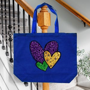 Puede incluir: Una bolsa de lona azul con un dise&ntilde;o de coraz&oacute;n de purpurina morado, dorado y verde. Los corazones est&aacute;n superpuestos.