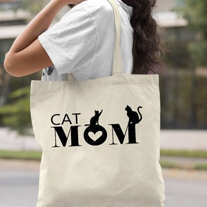 Puede incluir: Una bolsa de lona natural con texto negro y siluetas de gatos que dice "CAT MOM" con un coraz&oacute;n en el medio de la palabra "MOM".