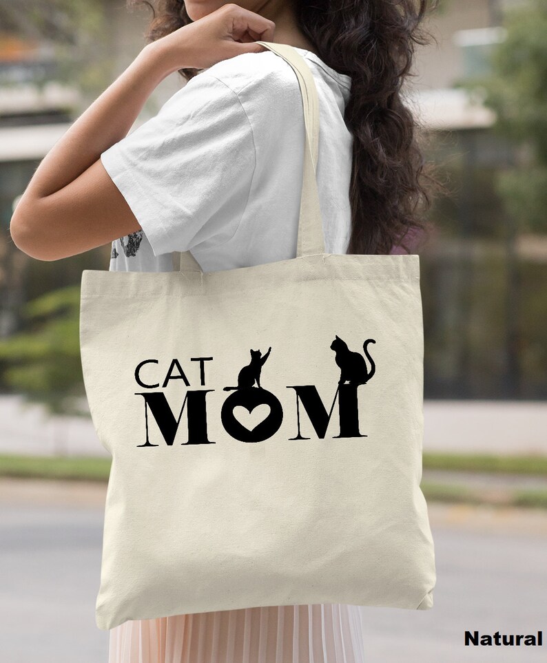 Puede incluir: Una bolsa de lona natural con texto negro y siluetas de gatos que dice "CAT MOM" con un coraz&oacute;n en el medio de la palabra "MOM".