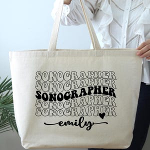 May include: A white canvas tote bag with the text "SONOGRAPHER" repeated in a retro font style. The name "emily" is written below the text with a small heart.