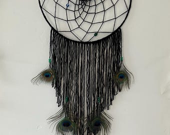 May include: A black dreamcatcher featuring a circular frame with a woven web pattern, accented by small colorful beads. Long black yarn strands hang from the bottom, interspersed with five peacock feathers and small green beads attached to the yarn.