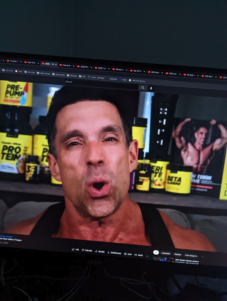 May include: A man with short dark hair is on a computer screen. The screen displays a YouTube video with the title "Mike O'Hearn". Behind him are several yellow and black supplement containers. A poster with a muscular man is visible.