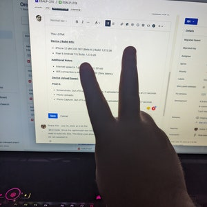 May include: A computer screen displays a software project with text including "ESALP-370" and "ESALP-278." The screen shows device information for an iPhone and Pixel phone. A hand is making a peace sign in front of the screen.