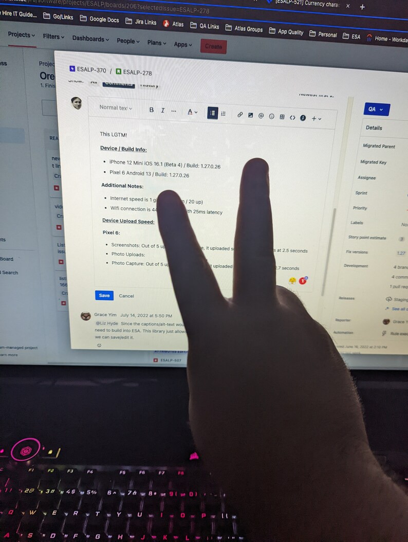 May include: A computer screen displays a software project with text including "ESALP-370" and "ESALP-278." The screen shows device information for an iPhone and Pixel phone. A hand is making a peace sign in front of the screen.