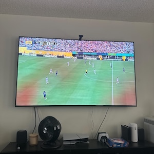 Peut inclure: Un grand écran plat affiche un match de football avec des joueurs en uniformes blancs et bleus sur un terrain vert. L'écran est monté sur un mur clair au-dessus d'un meuble de divertissement noir. Un ventilateur noir, des enceintes et des consoles de jeux sont visibles.