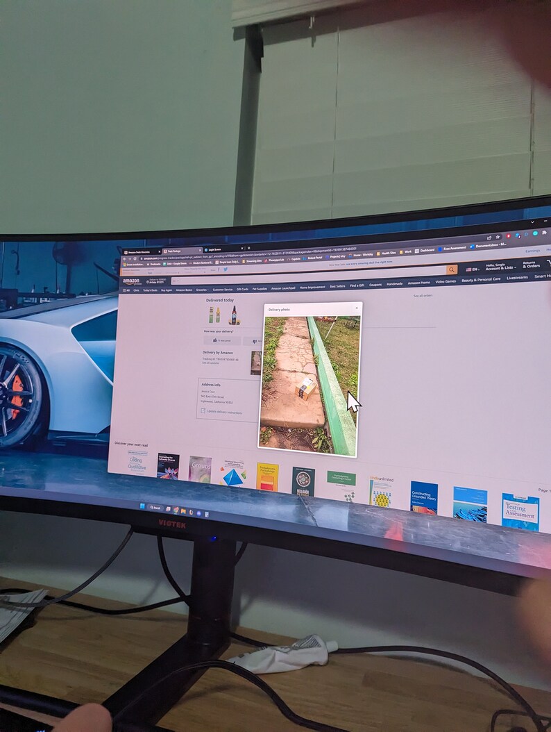May include: A computer monitor displaying a webpage with a delivery photo. The photo shows a small package on a brick pathway next to a green painted border. The monitor is on a wooden desk.