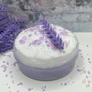 May include: A clear plastic jar filled with lavender-colored slime. The slime is topped with white foam, iridescent glitter, and lavender-colored sprinkles. A sprig of lavender sits atop the foam. Lavender sprigs are scattered around the jar.