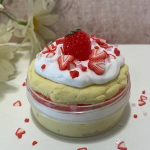 May include: A round clear plastic container filled with a pale yellow substance topped with white whipped cream, a single red strawberry, and small red strawberry-shaped slices. The container sits on a white surface with scattered red strawberry slices.