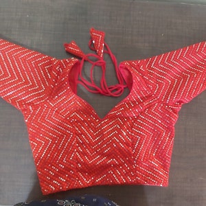 May include: A bright red blouse featuring a repeating chevron pattern created with small silver-toned sequins. The garment has short sleeves, a deep V-neckline, and red fabric ties at the back of the neck with matching sequined triangular accents.