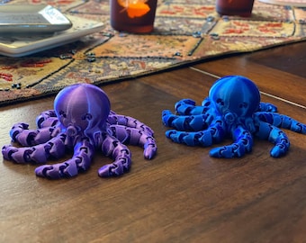 May include: Two 3D-printed octopus figurines. One is a gradient of purple hues, and the other is a gradient of blue hues. Each octopus has a rounded head and articulated tentacles, resting on a wooden surface.