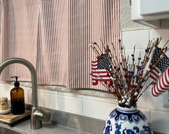 May include: Kitchen scene with red and white striped curtains hanging in front of a window. A blue and white floral vase holds a patriotic arrangement with American flags. A stainless steel faucet and soap dispenser are visible.