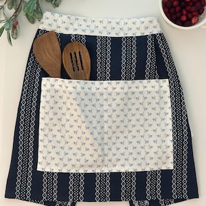 Op de afbeelding: Een marineblauwe schort met een witte zak en rand, met een blauw bloemmotief. Twee houten keukengerei zitten in de zak. Een kom met rode veenbessen staat op de achtergrond.