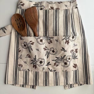 Puede incluir: Delantal color crema con rayas verticales negras y un bolsillo floral. El bolsillo presenta un patrón de flores blancas y detalles marrones. Dos utensilios de cocina de madera están en el bolsillo.