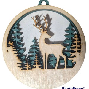 May include: A wooden Christmas ornament featuring a deer silhouette standing in a forest scene. The deer is cut out of the wood, and the forest is painted in shades of green and blue.