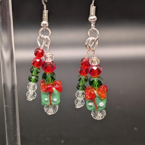 May include: A pair of festive earrings featuring red, green, and clear beads. Each earring has a green gift box charm with a red bow and white accents, suspended from silver-tone hooks.