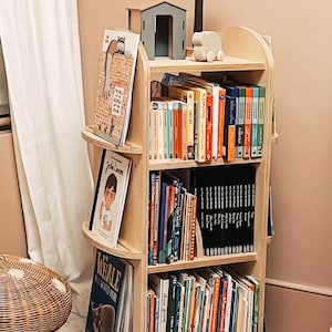 May include: A wooden bookshelf with rounded edges and shelves filled with colorful children's books. The bookshelf is in a room with a light pink wall and a white curtain.