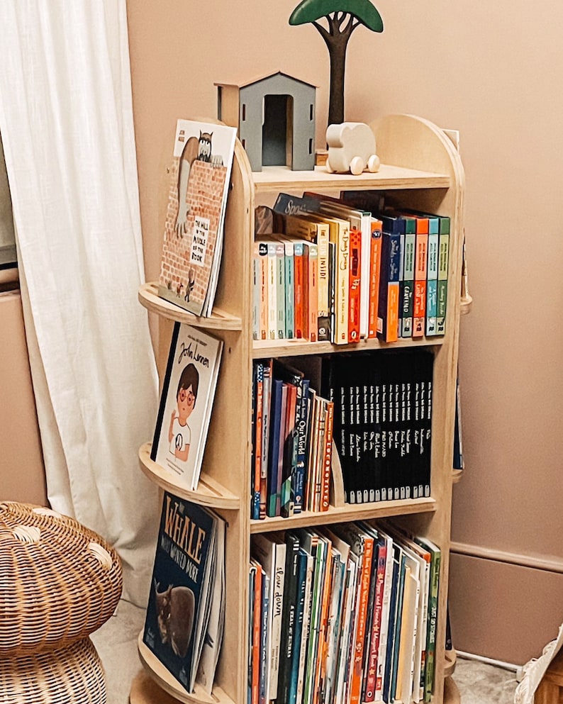 May include: A wooden bookshelf with rounded edges and shelves filled with colorful children's books. The bookshelf is in a room with a light pink wall and a white curtain.