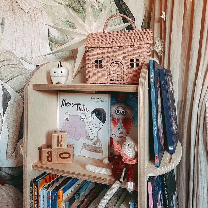 May include: A wooden bookshelf with multiple shelves, filled with books and toys. The top shelf has a wicker house-shaped basket, a white ceramic figurine, and a star-shaped decoration. The middle shelf has a children's book titled "Mein Tutu" and wooden blocks with letters. The bottom shelf has a variety of books, including one with the title "Die Kleine Fledermaus".