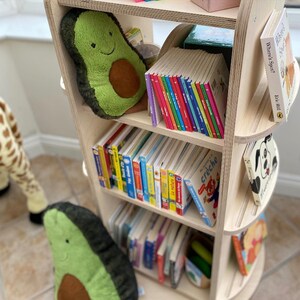 May include: A wooden bookshelf with rounded edges features multiple shelves filled with colorful children's books. The top shelf has a plush avocado-shaped toy and a stack of colorful board books. The bookshelf is a great addition to a child's room.