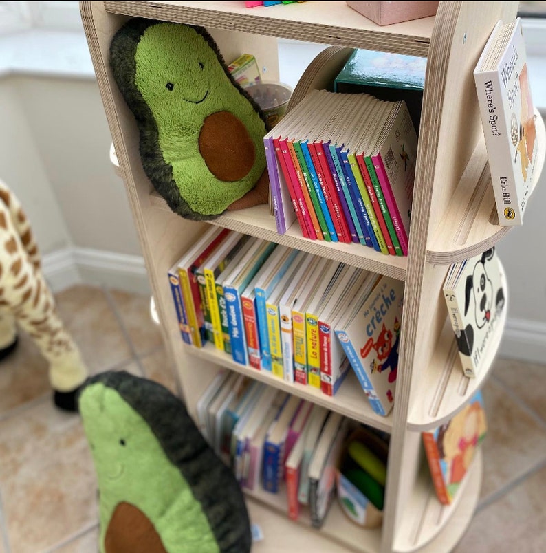 May include: A wooden bookshelf with rounded edges features multiple shelves filled with colorful children's books. The top shelf has a plush avocado-shaped toy and a stack of colorful board books. The bookshelf is a great addition to a child's room.
