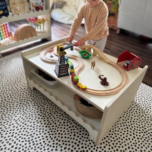 May include: A white wooden play table with a train track set up on top. A child is playing with the train set. The table has a shelf underneath. The table is sitting on a black and white polka dot rug.