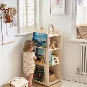 May include: A light wood rotating bookshelf with a child standing in front of it. The bookshelf has five shelves and is filled with books and toys. The child is wearing a white and yellow floral print onesie. The bookshelf is on a white rug.
