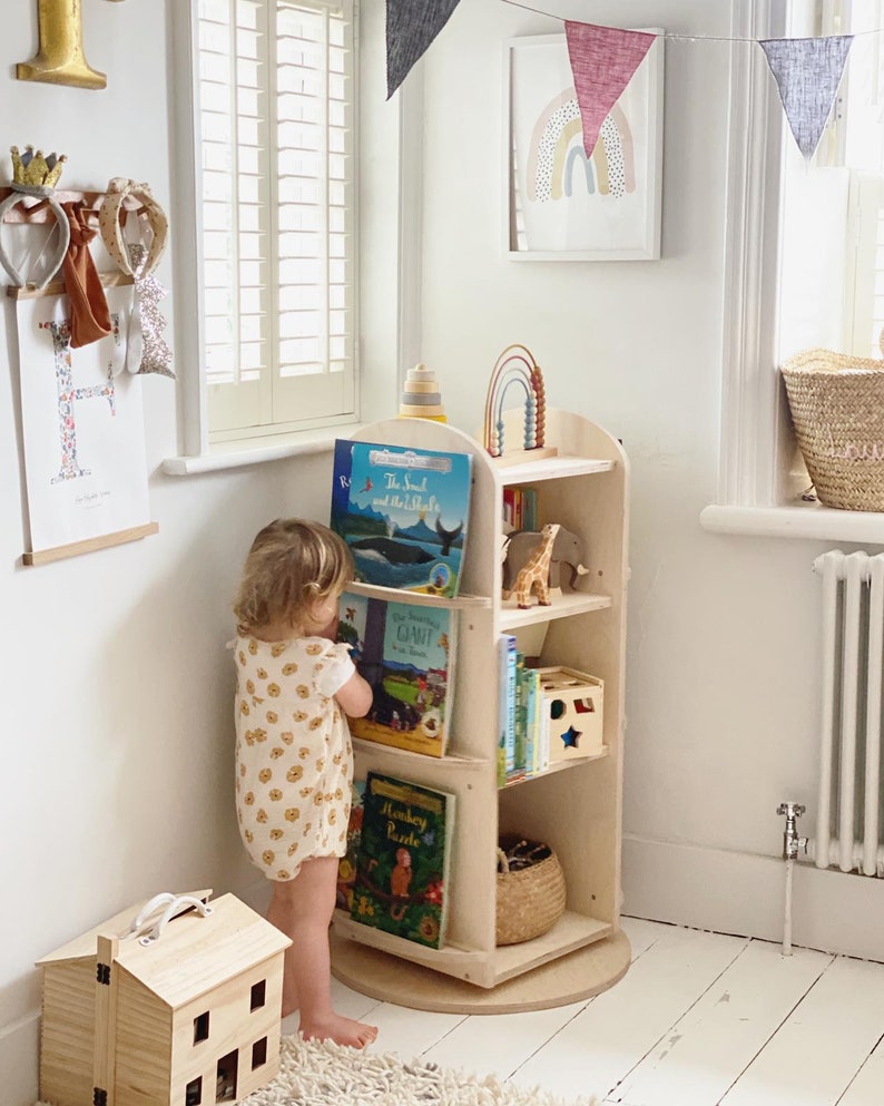May include: A light wood rotating bookshelf with a child standing in front of it. The bookshelf has five shelves and is filled with books and toys. The child is wearing a white and yellow floral print onesie. The bookshelf is on a white rug.