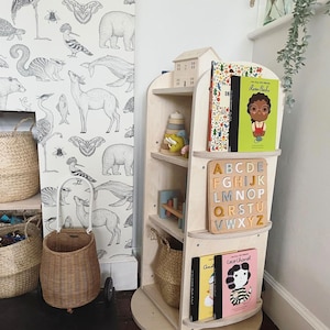 May include: A wooden rotating bookshelf with four shelves. The shelves are filled with books and toys. The bookshelf is on a white base and has a white background. The bookshelf is in a room with a white wall and a black and white wallpaper.