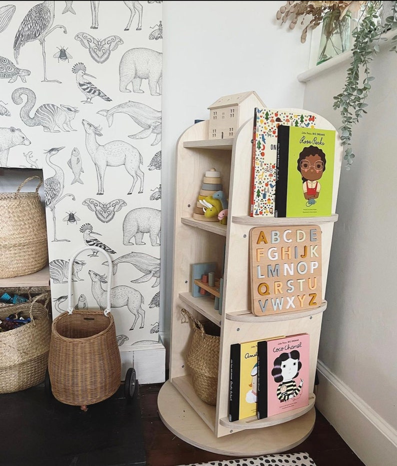 May include: A wooden rotating bookshelf with four shelves. The shelves are filled with books and toys. The bookshelf is on a white base and has a white background. The bookshelf is in a room with a white wall and a black and white wallpaper.