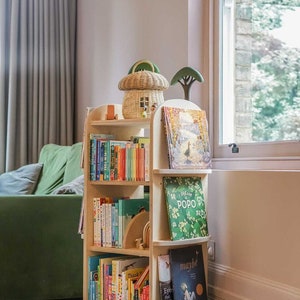 May include: A wooden rotating bookshelf with multiple shelves filled with colorful children's books. The bookshelf is in a room with a green couch and a window with a view of trees.