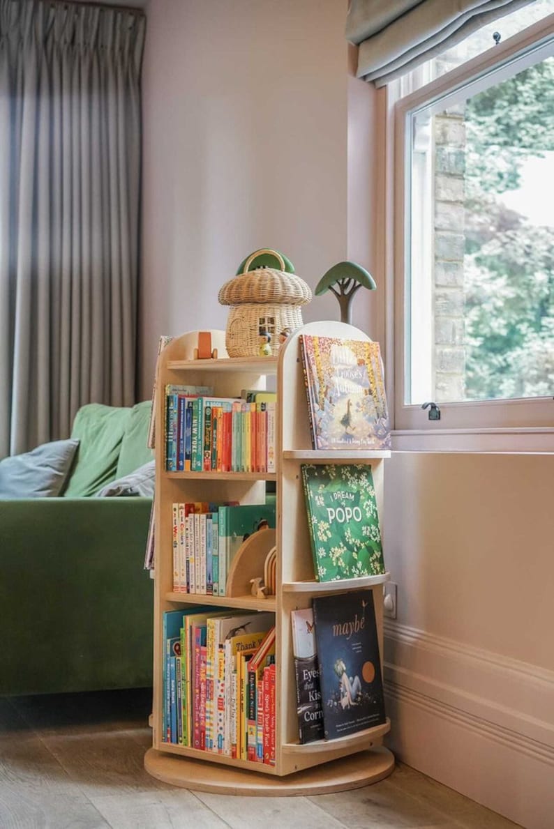 May include: A wooden rotating bookshelf with multiple shelves filled with colorful children's books. The bookshelf is in a room with a green couch and a window with a view of trees.