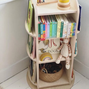 May include: A wooden rotating bookshelf with three shelves, filled with colorful children's books. The top shelf has a wooden abacus and a wooden stacking toy. The bottom shelf has a woven basket and a stuffed giraffe toy.